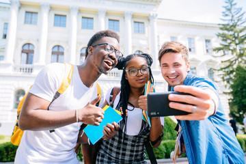Three multiracial friends having fun and joking together making common selfie for social networks, hipster girl and two boys in casual wear taking photos on smartphone enjoying time together.