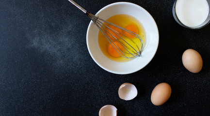 Ingredients for omelette, fresh chicken eggs, eggs and yolks in a white bowl, milk, a whisk. Black background, top view, copy space