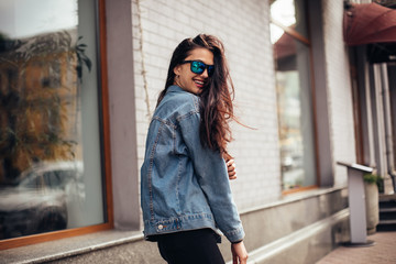 Obraz premium Happy attractive young woman in sunglasses walking in the city. Playful woman wraps herself over her shoulder and looking in camera.