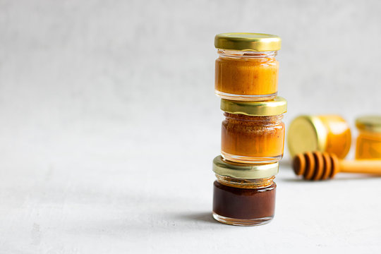 Three Small glass jar with metal cap with different kinds and colours of honey one on another and spoon isolate and on grey cement background with copy space. Healthy product, natural. Horizontal