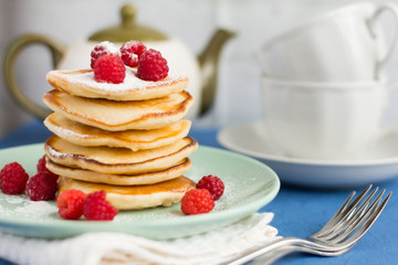stack of baked homemade pancakes with raspberries, sprinkled with powdered sugar and tea utensils in the background, selective focus