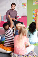 Elementary Pupils Raising Hands To Answer Question As Male Teacher Reads Story In Classroom