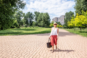 Fototapeta na wymiar Beautiful young positive and independent French hipster girl walking in the park in the city with hat and sunglasses in retro or vintage fashion clothes holding the bag in her hand