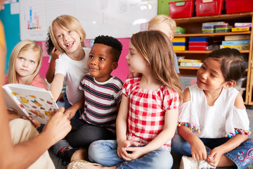 Group Of Elementary School Pupils Sitting On Floor Listening To Female Teacher Read Story