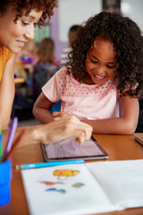 Elementary School Teacher And Female Pupil Drawing Using Digital Tablet In Classroom