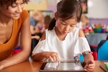 Elementary School Teacher And Female Pupil Drawing Using Digital Tablet In Classroom