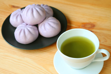 Closeup a cup of green tea with blurry pile of purple sweet potato steamed buns on wooden table