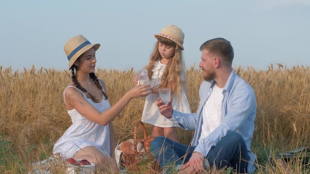 family idyll, young happy couple with little nice child girl drink milk at weekend countyside picnic in sunny crop season of yellow grain meadow against blue sky