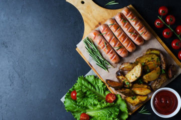 Grilled sausages on a wooden chopping Board. Fried potatoes, rosemary, tomatoes, green lettuce leaves, tomato ketchup. Unhealthy diet. Dark background. Copy space