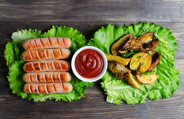 Grilled sausages on green lettuce leaves, tomato sauce, red cherry tomatoes on a branch. Wooden dark background