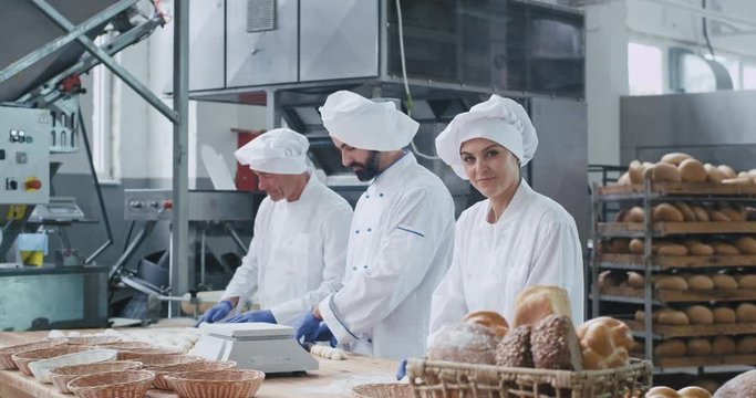 Beautiful bakery factory stylish bakers two men and one woman working together forming pieces of dough for baking bread looking to the camera and smiling large