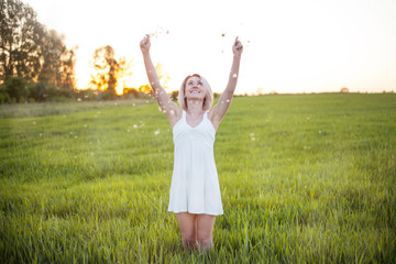 happy girl in the field. The blonde girl in a tiny white dress with dandelions happily smiles looking up. Green field. Happy moment