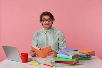 Young smiling man in glasses wears in shirt, student sits by the table and working with notebook, prepared for exam, reads book, having funny look, isolated over pink background.