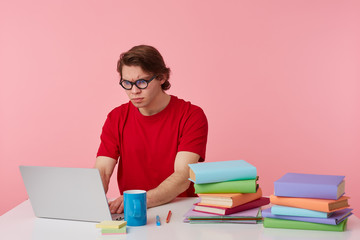 Frowning young student in glasses wears in red t-shirt, man sits by the table and working with laptop and books, looks at the monitor with surpraised, something wrong, isolated over pink background.