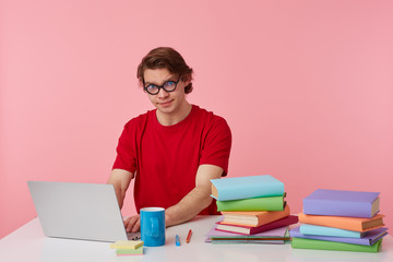 Photo of young student in glasses wears in red t-shirt, man sits by the table and working with laptop and books, isolated over pink background. Looks displeased and unhappy.