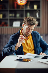 Young serious bearded Caucasian blogger dressed smart casual writing notes in agenda while sitting...