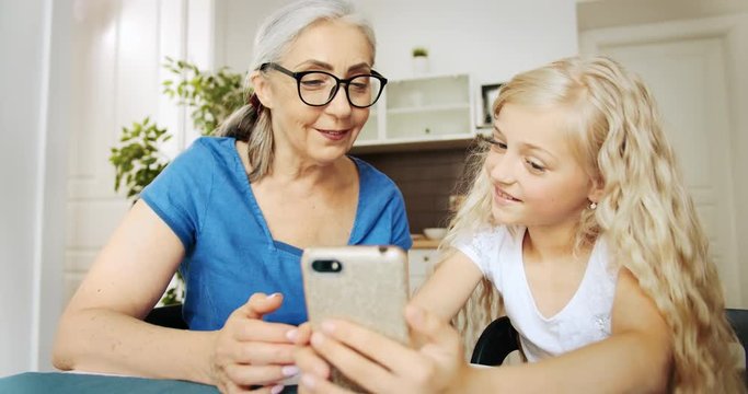 Happy girl in white blouse sitting with granny in kitchen and scrolling smartphone, showing photos in application