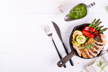 Chicken breast grill with bbq vegetables and pesto sauce in a cast iron pan on a white background, flat lay,