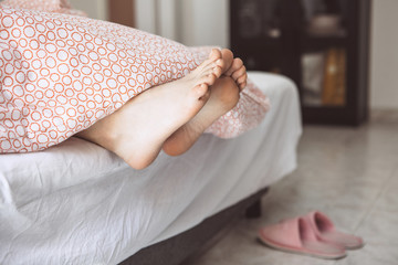 Legs of young adult girl under the blanket close up. She is sleeping. Near slippers on the floor