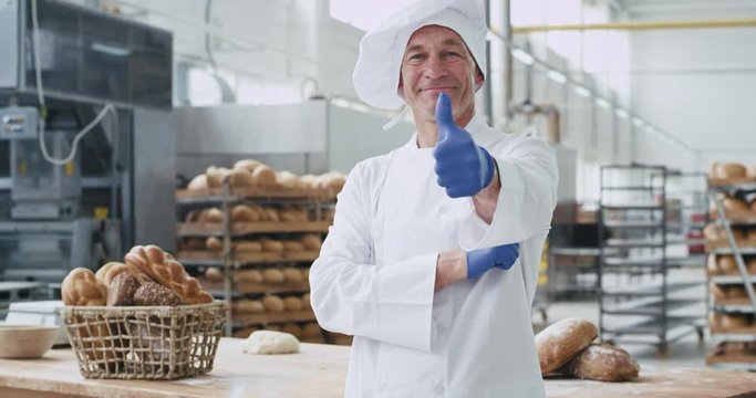 Charismatic old baker man in a stylish uniform looking straight to the camera and smiling at bakery industry showing big like