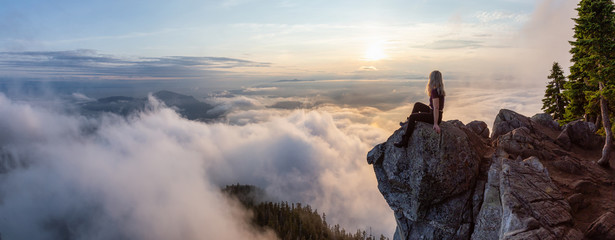 Adventurous Female Hiker on top of a mountain covered in clouds during a vibrant summer sunset....