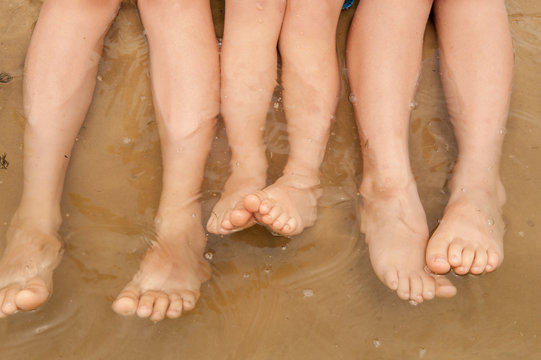 Funny kids in bathing suits sit together on sand with their legs in water. boys are happy on holiday in village together. Summer day, river, swimming in the water. Close-up legs and feet