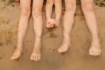 Funny kids in bathing suits sit together on sand with their legs in water. boys are happy on holiday in village together. Summer day, river, swimming in the water. Close-up legs and feet