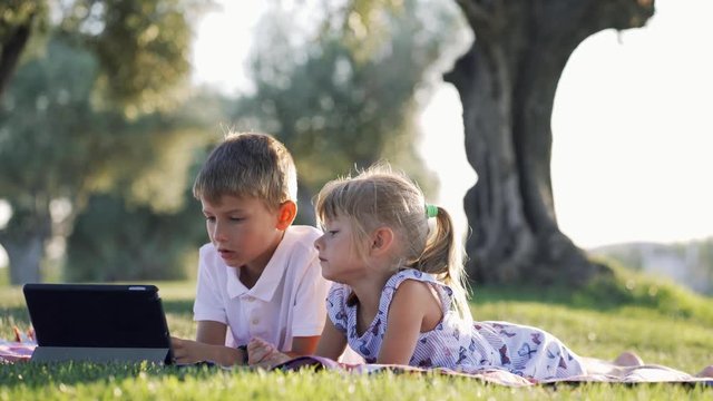 boy and girl brother and sister use the tablet in the summer in the park on vacation watching cartoons movies series smiling and happy
