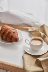 fresh croissant on plate with coffee in cup on wooden tray in bed at morning