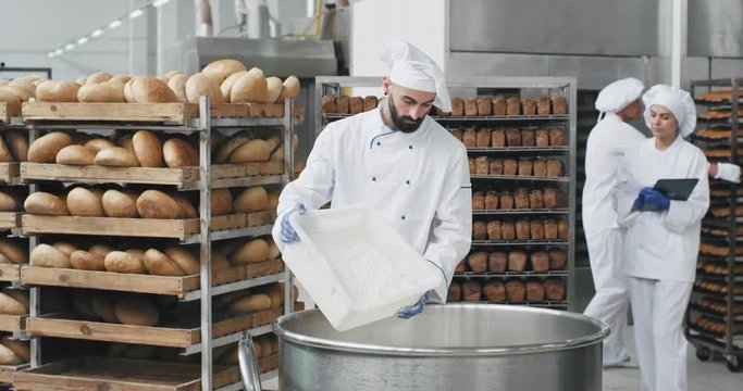 Bakery industry big chef in a special uniform preparing the dough add some flour from the basket bakery engineer monitoring all the process background baked bread on the shelves. slow motions
