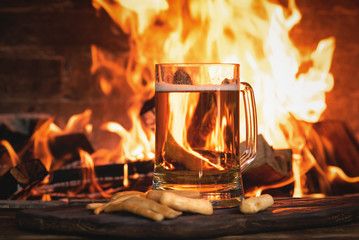 Beer in a mug and cheese sticks on a wooden table on a burning fire in a fireplace background.