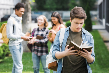selective focus of teenager reading book with friends on background