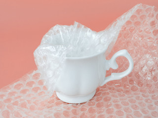 White elegant tea cup half packaged with white transparent bubble wrap on a pink background....