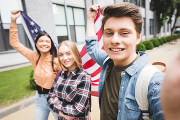 smiling teenagers taking selfie and holding american flag outside