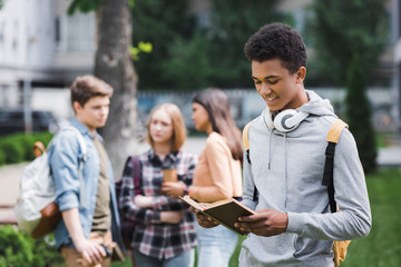 smiling african american teenager in hoodie with headphones reading book