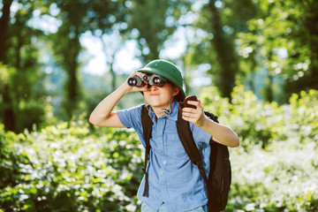 Portrait of little explorer with binoculars and  compass in forest. Boy traveler in helmet play in the park. Happy child go hiking with backpack in summer nature.