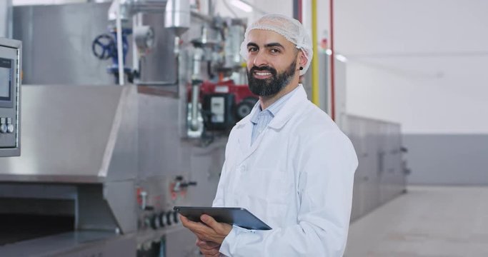 Stylish engineer man with a beard in a special white uniform monitoring the industrial machine