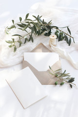 Summer wedding stationery mock-up scene. Blank greeting cards, envelope, olive branches and silk ribbon. White background with cotton table runner in sunlight, shadows. Flat lay, top view, vertical