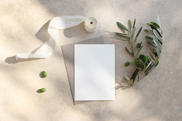 Summer wedding stationery mock-up scene. Blank greeting card, invitation. Craft envelope, olive fruit, branch and silk ribbon. Elegant marble background in sunlight, shadows. Flat lay, top view.