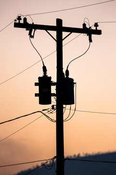 An silhouetted electricity transformer on a power pole at sunset.