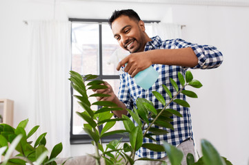 people, nature and plants care concept - smiling indian man spraying houseplant by water sprayer at...