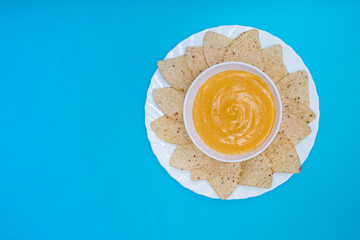 Queso cheddar vegano en un bol con nachos para mojar sobre un fondo azul.