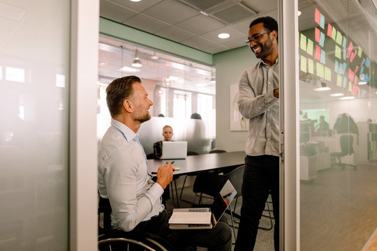 Smiling businessman looking at disabled colleague sitting on wheelchair in board room