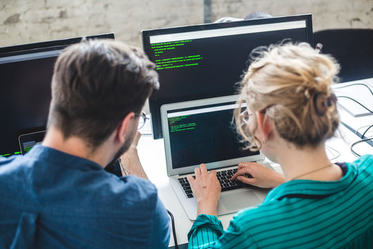 Rear view of female IT expert coding in laptop while sitting by male hacker at workplace