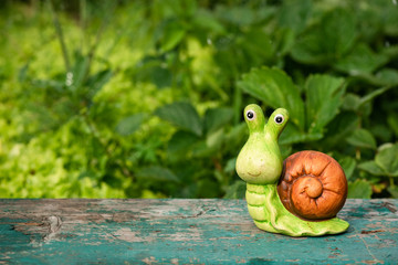 A toy snail on a wooden board on a green garden flower bed background with copy space.