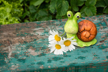 A toy snail on a wooden board on a green garden flower bed background with copy space.