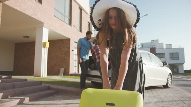 portrait of a pretty girl in summer hat moving a suitcase to the car. happy parents loading luggage into a trunk preparing for a summer vacation and roadtrip.