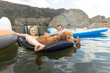 Two teenage girls and a golden retriever dog lounge comfortably on inflatables at a lake
