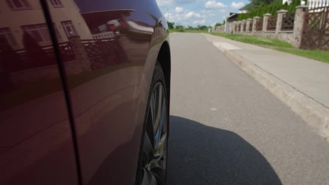 Front tire of glossy red car spinning, wheel of automobile riding on asphalt road, point of view. Auto driving through cottage town, modern suburban houses reflected on glossy car surface