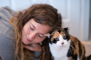 Closeup portrait of happy smiling young woman bonding with calico cat pet companion, bumping rubbing bunting heads, friends showing affection